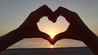 Zwei Hände formen ein Herz und lassen die untergehende Sonne durchscheinen